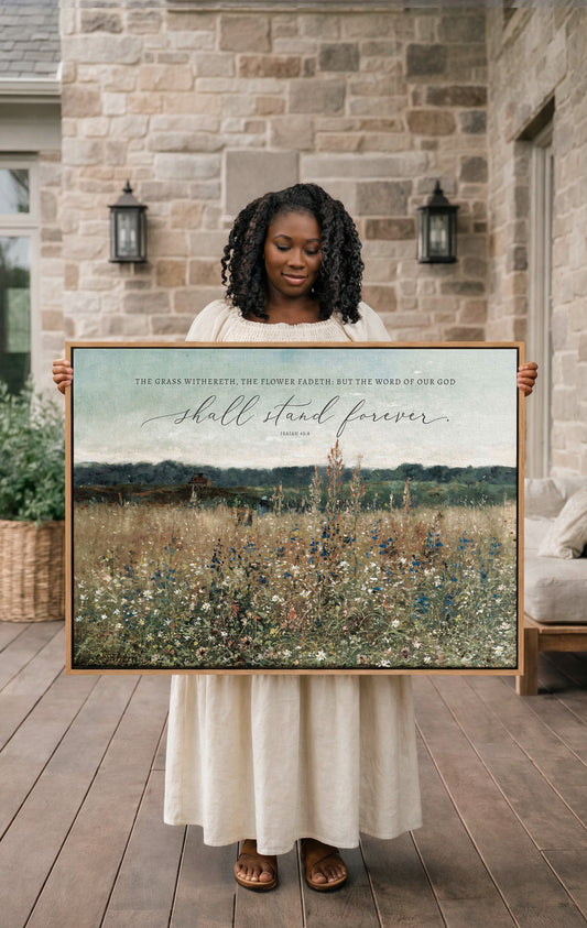 A woman in a white dress stands holding a large framed print of a field with flowers, displaying a Bible verse.