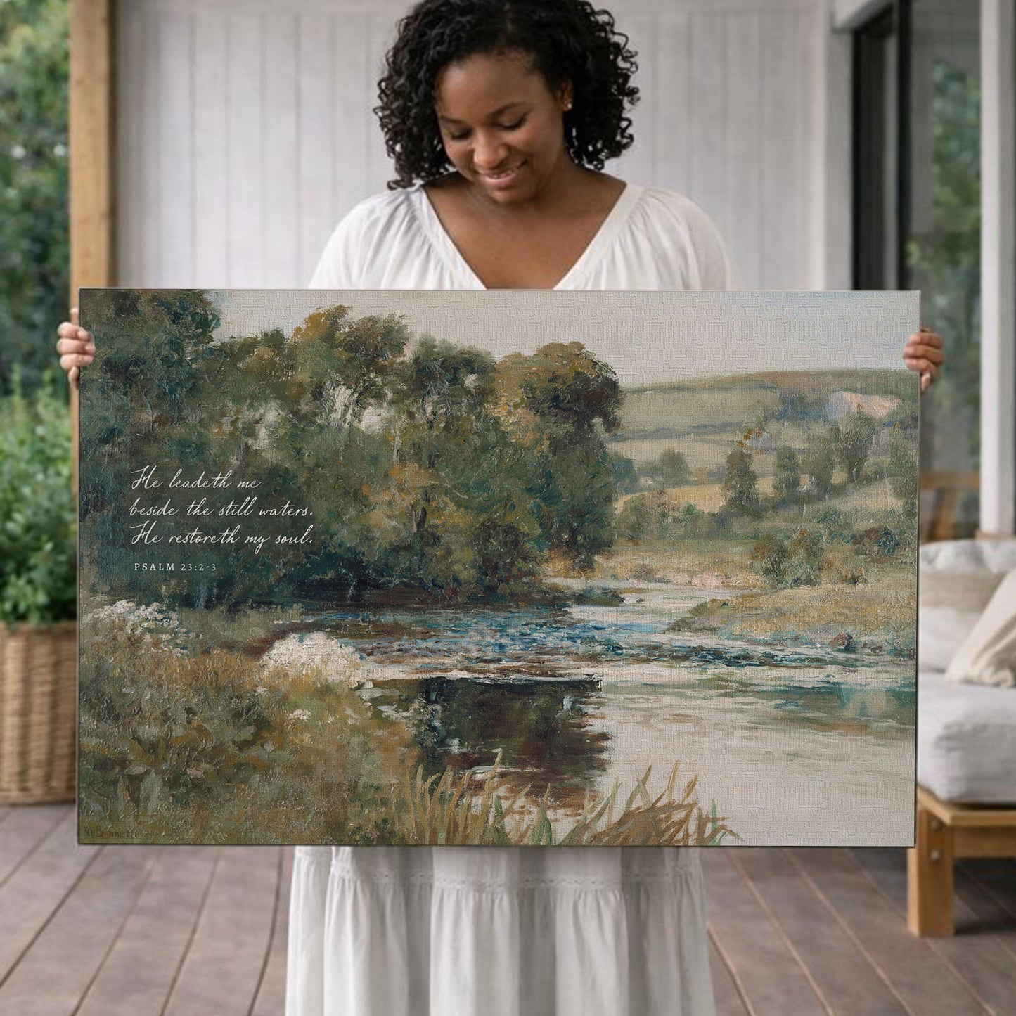 A woman holds a large painting of a serene landscape with a river and trees, featuring a Bible verse from Psalm 23:1.