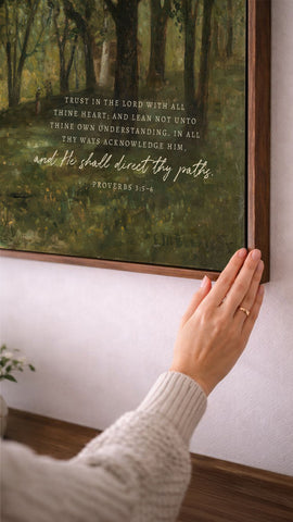 A person's hand is reaching towards a framed painting of a forest scene, with a Bible verse visible on the painting.