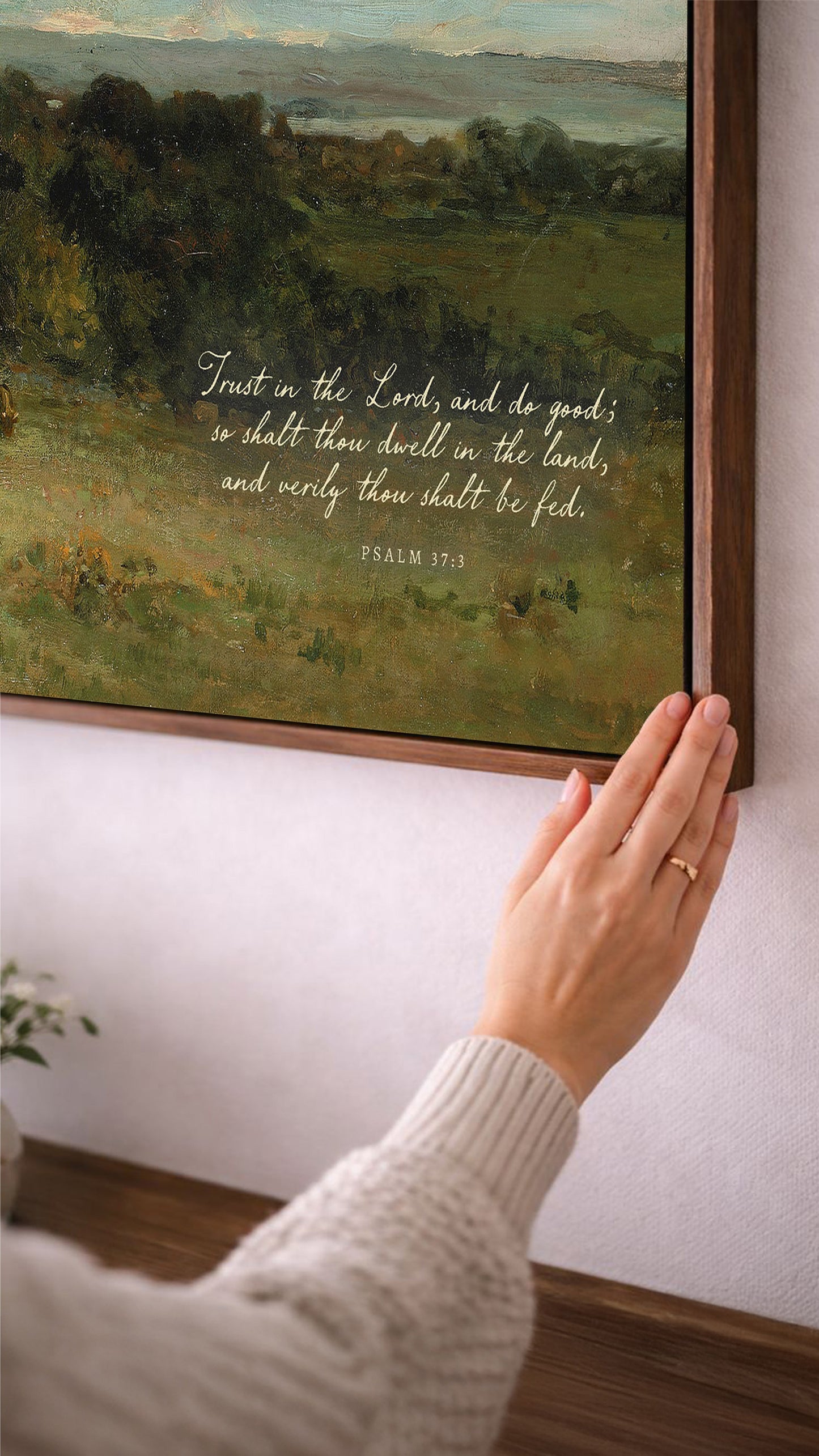 A person's hand is reaching towards a framed painting of a pastoral landscape, with a Bible verse visible in the painting.
