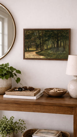 a wooden console table with various items on it, including a framed landscape painting, a lamp, a bowl, and a plant. The table is set against a white wall, and there is a round mirror above it.