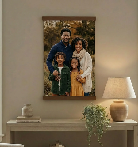 a family portrait hanging on a wall, with a wooden table below it. The table has a lamp and a plant on it. There are also some books and a vase on the table.