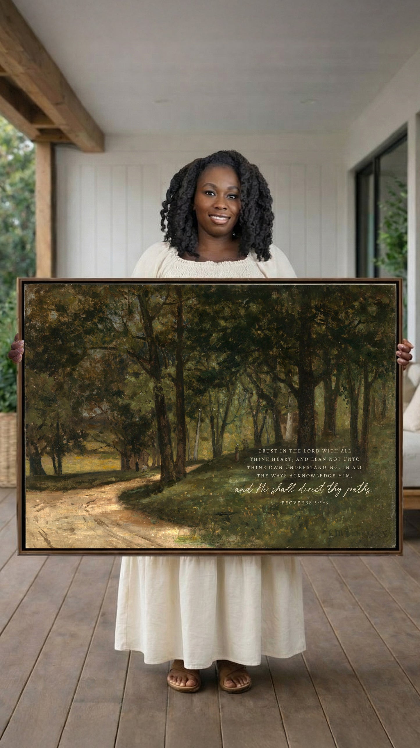 A woman in a white dress holds a large painting of a forest scene, smiling and looking directly at the camera.