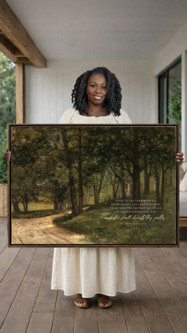 A woman in a white dress holds a large painting of a forest scene, smiling and looking directly at the camera.