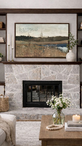 a cozy living room with a stone fireplace, a framed landscape painting above it, and a wooden coffee table with a vase of white flowers.