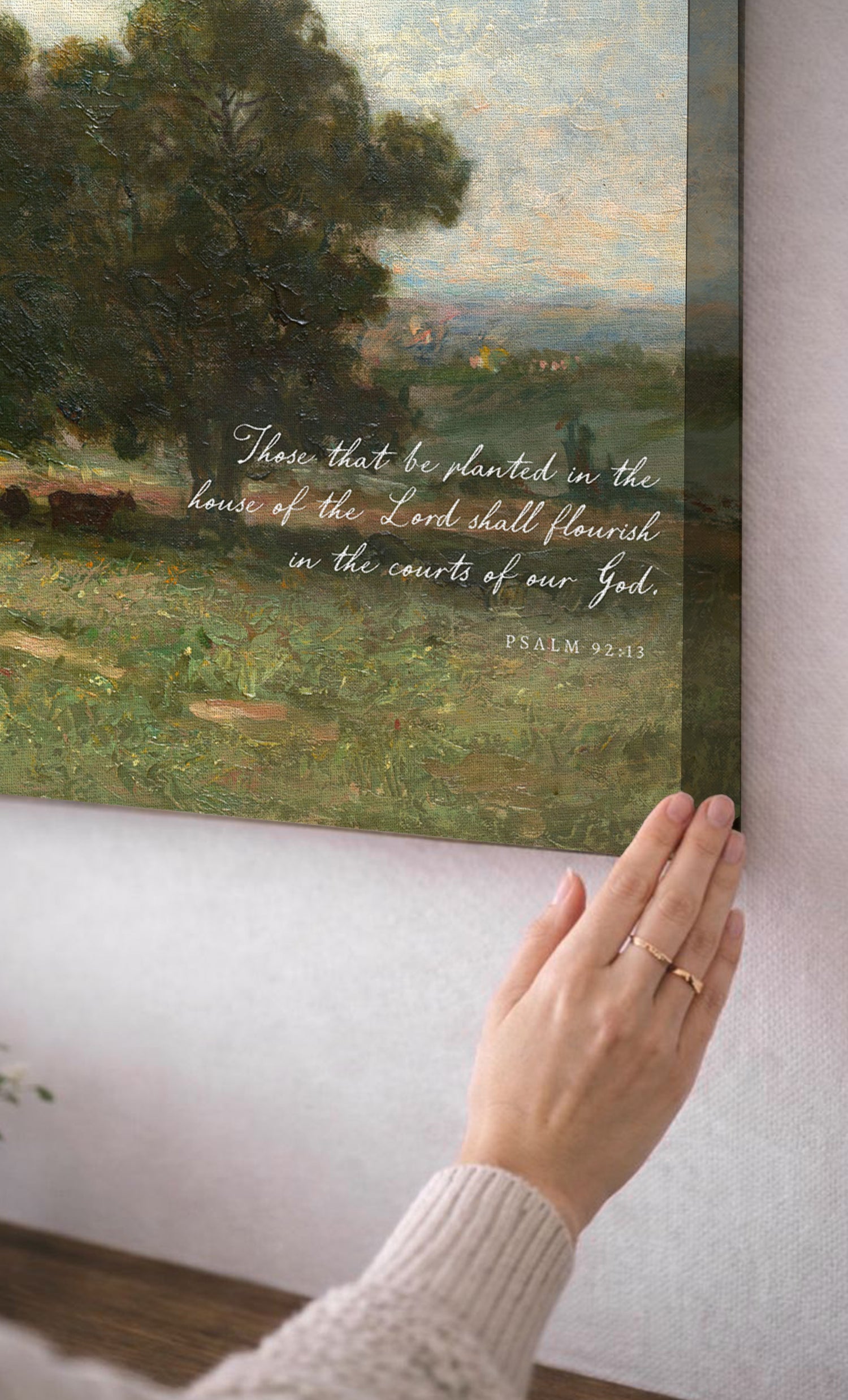 A person's hand is holding up a framed painting of a pastoral landscape, with a text overlay that reads "These that be planted in the house of the Lord shall flourish in the courts of our God."